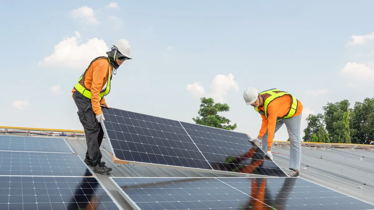 Zwei Monteure in Warnwesten und Helm setzen ein Photovoltaikmodul auf einem Gewerbedach mit Trapezblech ein