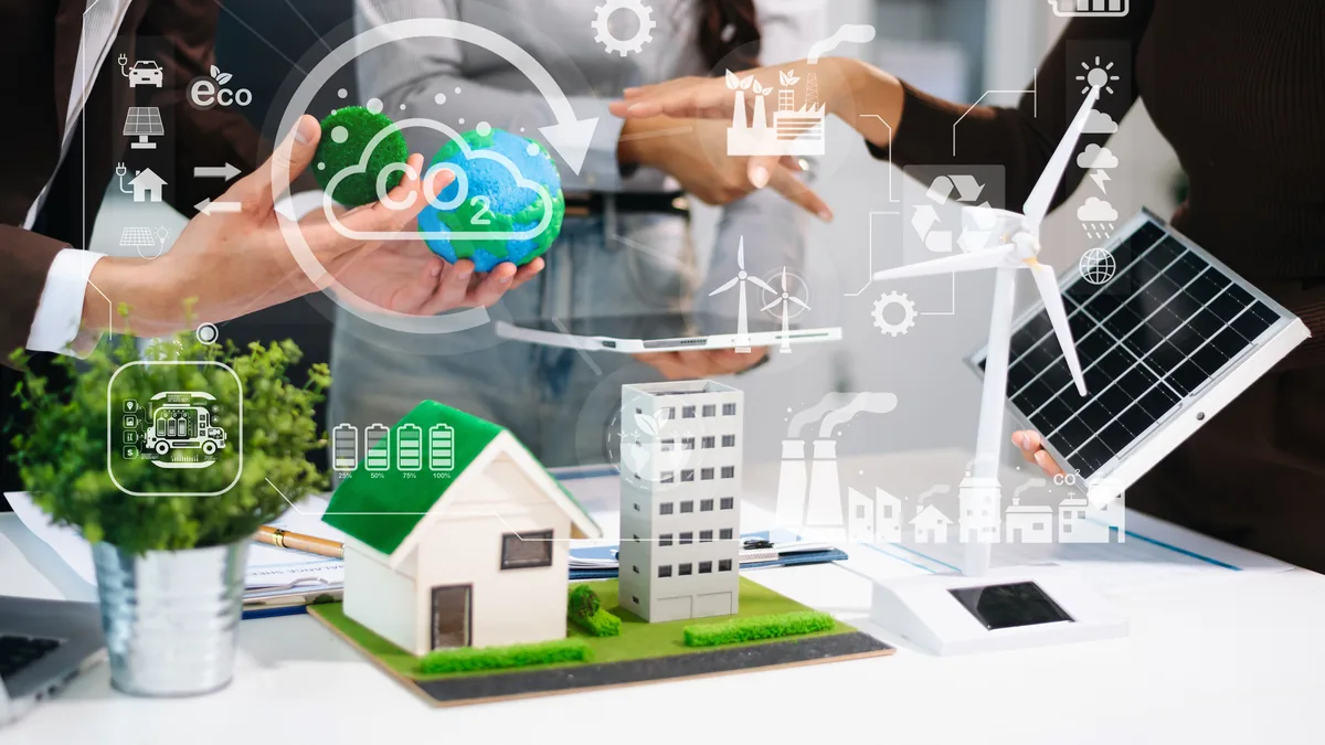 Haus- und Gebäudemodell, Windrad und Solarpanel auf einem Schreibtisch, darüber halten Hände ein Globusmodell mit CO2-Symbol